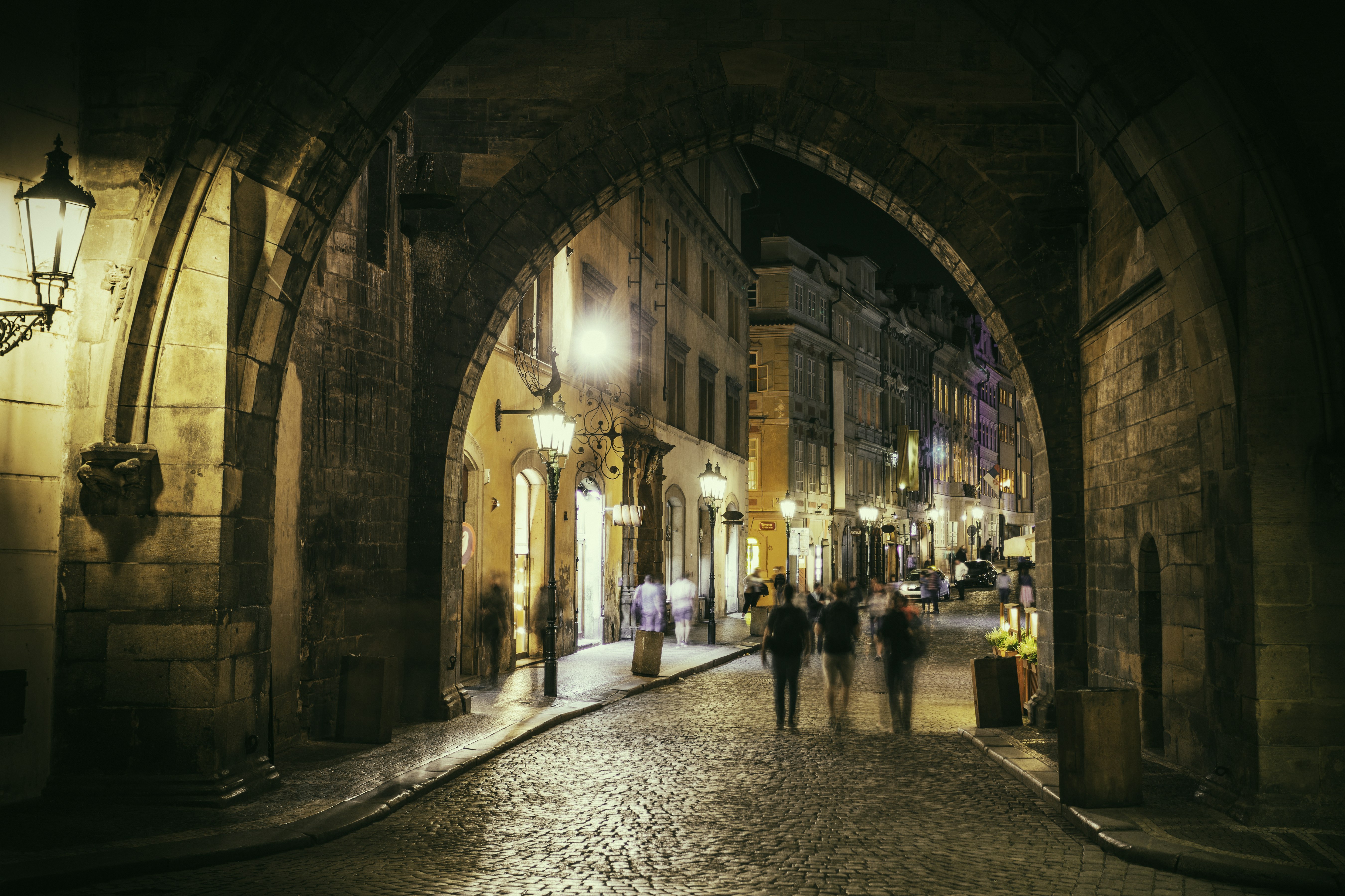 A group of people walk down a cobbled stone street towards a busy Prague street at night
